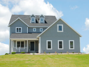 Two-story blue house with gable roof and dormer windows on a green lawn under a blue sky